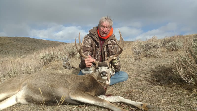 A hunter kneels behind a harvested mule deer in a field.