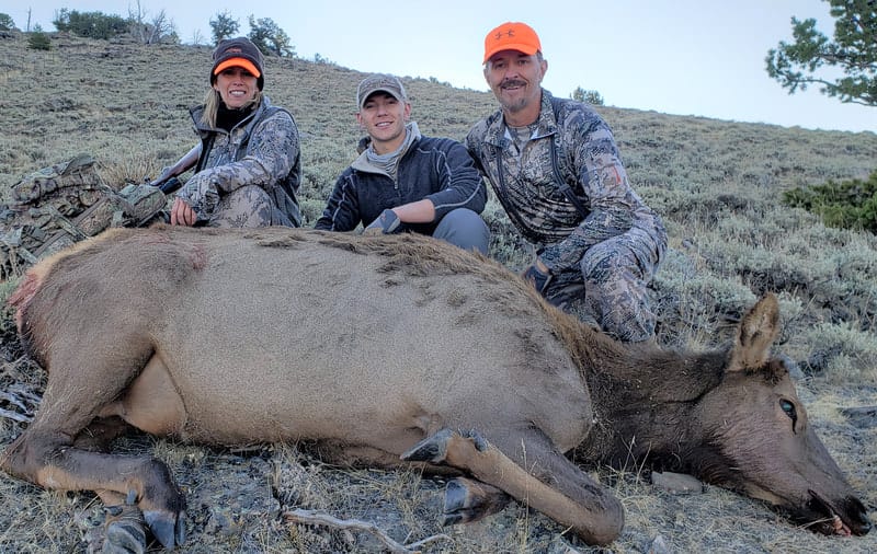 Three hunters pose with a harvested elk on a hillside.