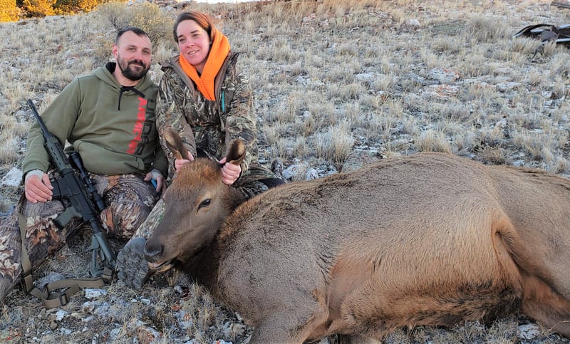 Two hunters pose with a harvested cow elk in a dry, grassy field.
