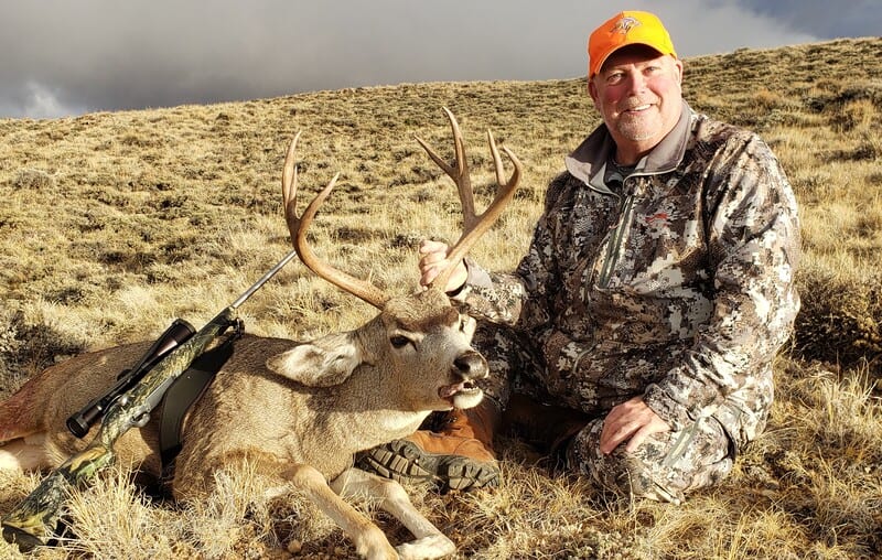 A hunter in camouflage poses with a harvested mule deer in a dry, grassy field.