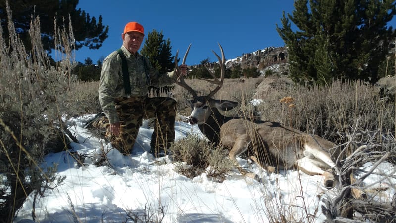 A hunter kneels next to a harvested mule deer in a snowy, brushy landscape.