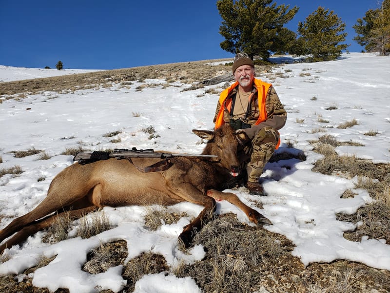 A hunter poses with a harvested elk in a snowy, mountainous landscape.
