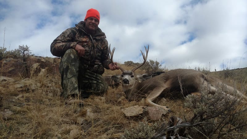 A hunter kneels next to a harvested mule deer on a grassy hillside under a cloudy sky.
