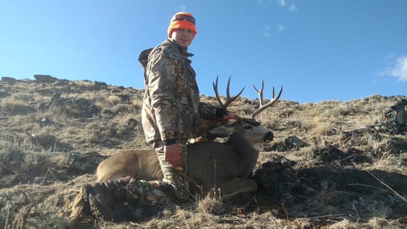 A hunter in camouflage and an orange hat poses with a harvested mule deer on a rocky, grassy hillside under a blue sky.