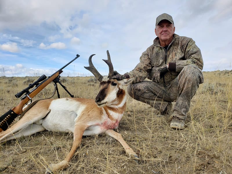 A hunter kneels next to a harvested pronghorn with a rifle on a tripod in a grassy field under a cloudy sky.