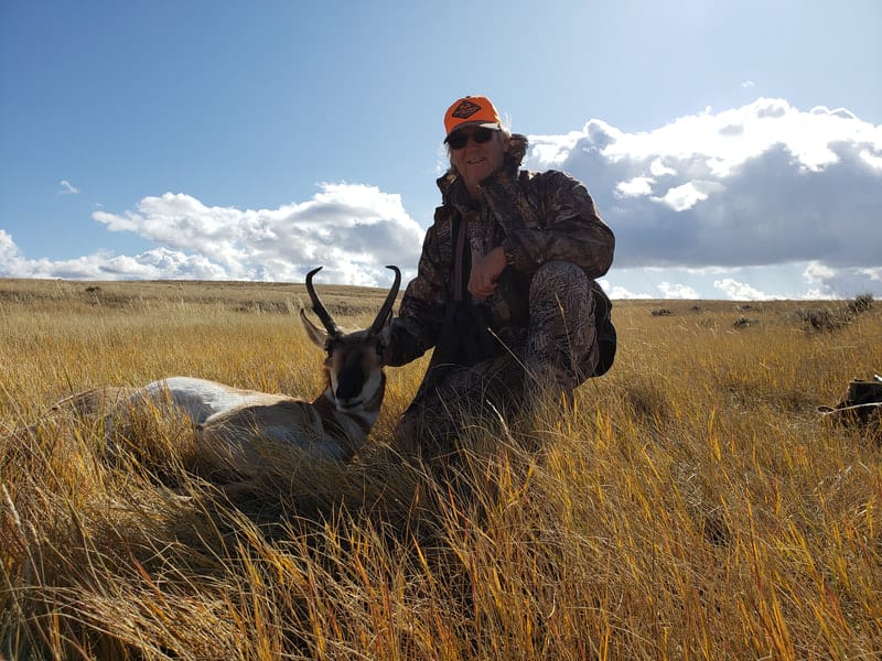 A hunter kneels next to a harvested pronghorn in a grassy field under a blue sky.