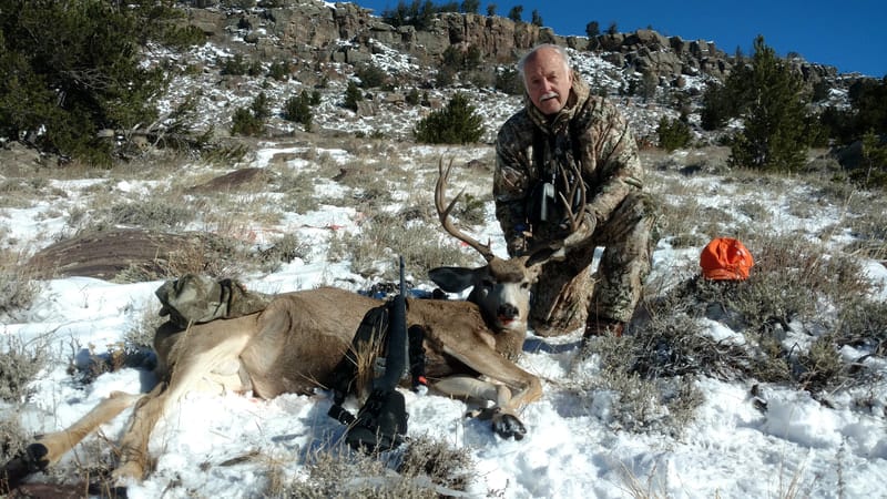 A hunter kneels next to a harvested mule deer in a snowy, rocky landscape.