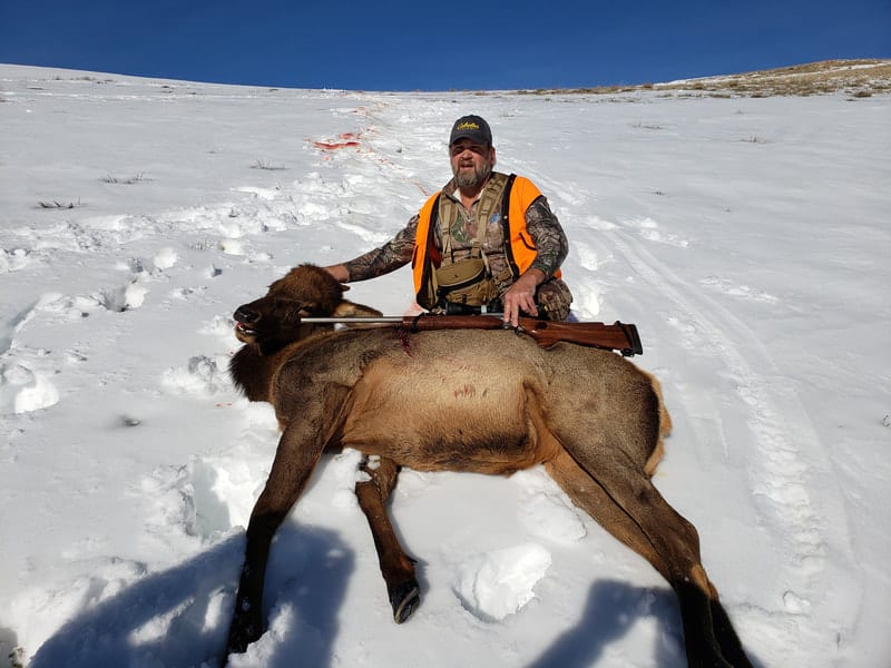 A hunter poses with a harvested elk in a snowy landscape.