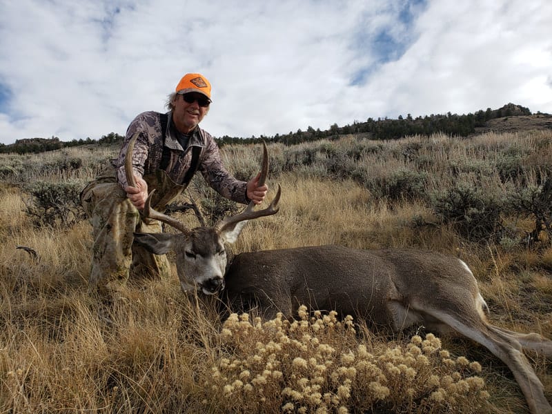 A hunter poses with a harvested mule deer in a grassy field.