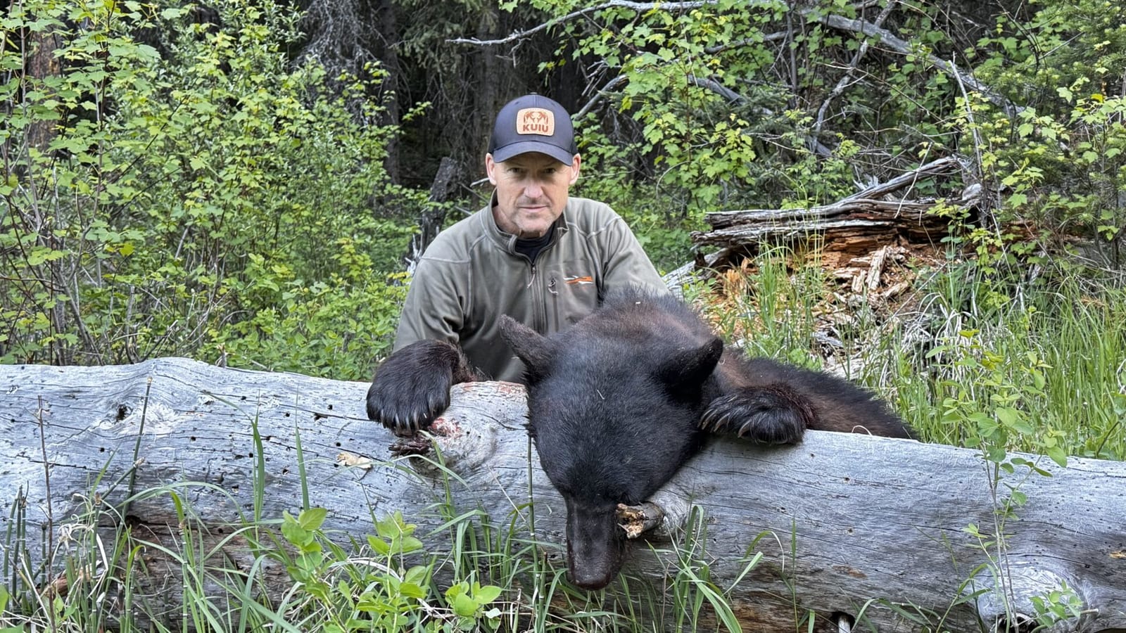 A hunter poses with a harvested black bear next to a fallen log in a forest.