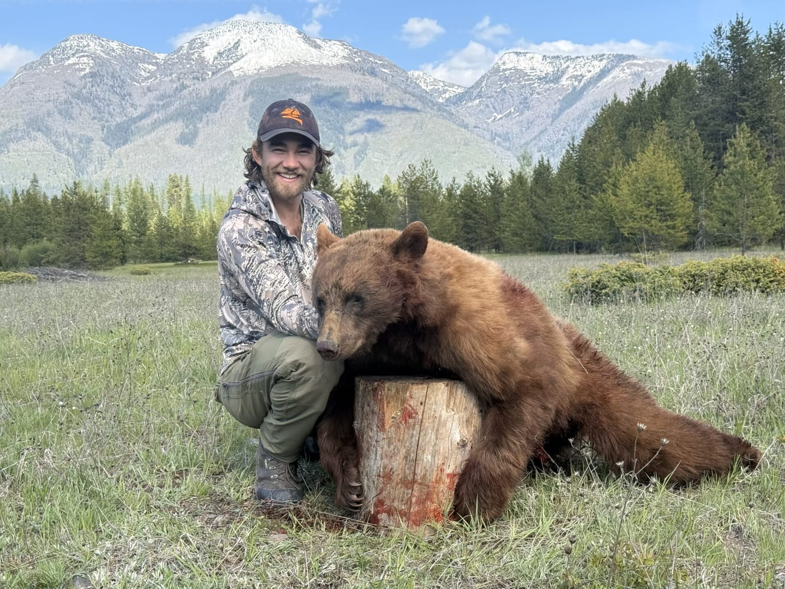 A hunter poses with a harvested black bear in a grassy field with mountains in the background.