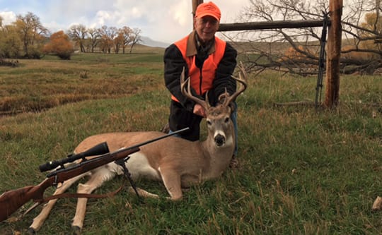 A hunter in an orange vest poses with a harvested whitetail deer and a rifle in a grassy field.