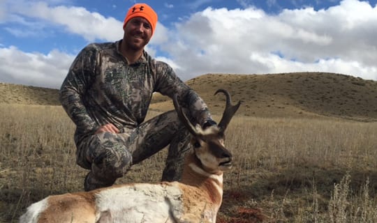 A hunter in camouflage kneels next to a harvested pronghorn in a dry, grassy field under a cloudy sky.
