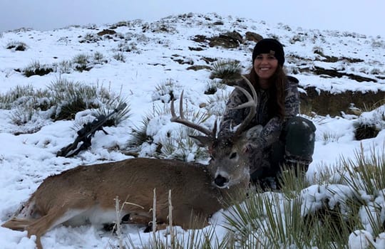 A woman in camouflage poses with a harvested mule deer in a snowy, grassy field.