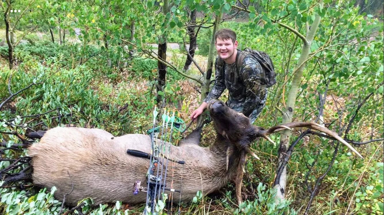 A hunter in camouflage poses with a harvested elk in a forest.