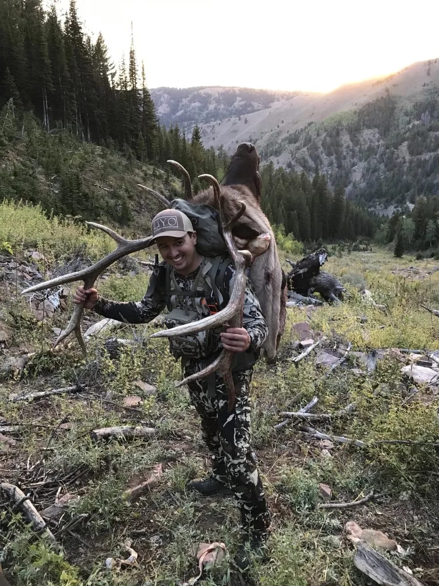 A hunter smiles while packing out a harvested elk with antlers in a mountainous, forested area.