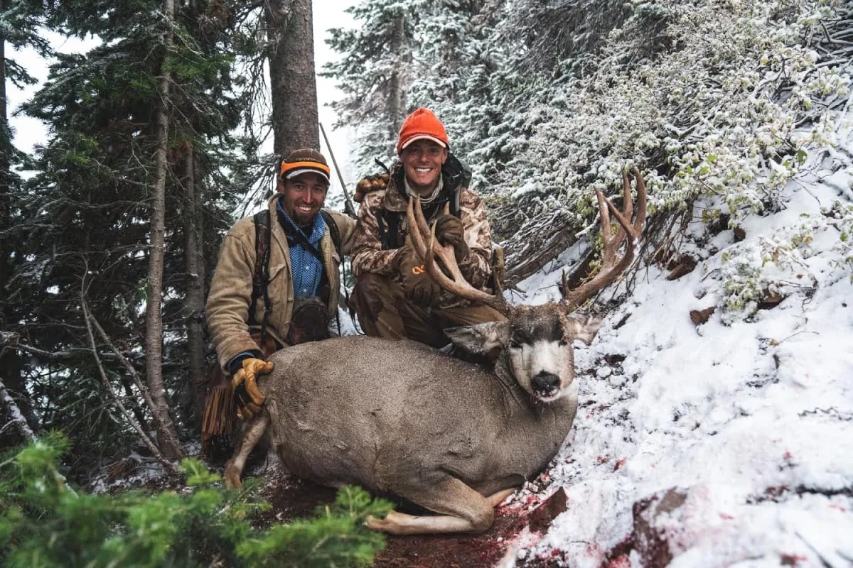 Two hunters pose with a harvested mule deer in a snowy forest.