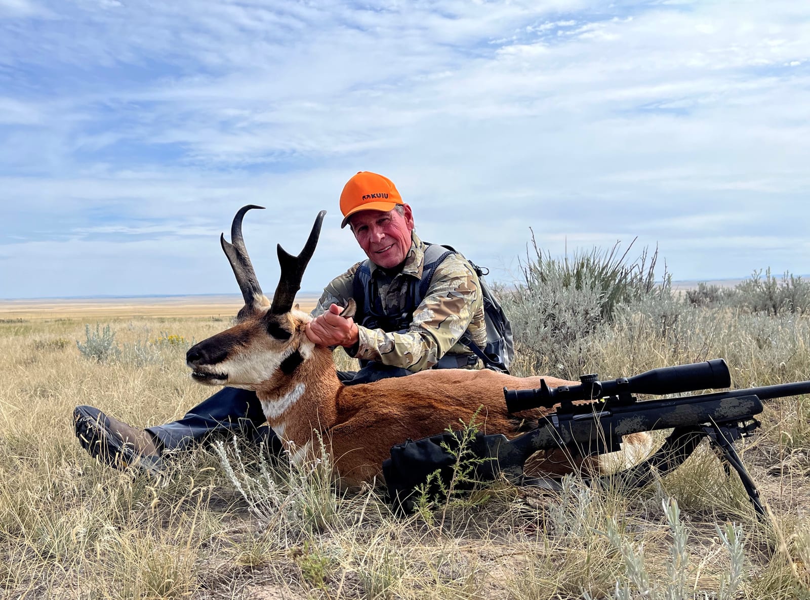 A hunter in an orange hat poses with a harvested pronghorn in a grassy field.