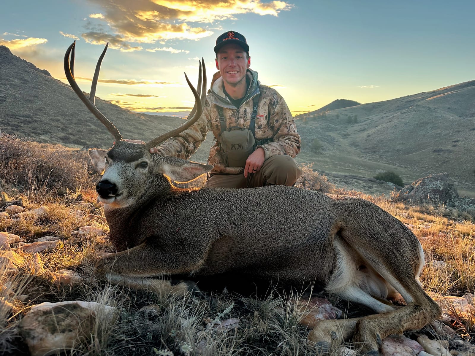 A hunter poses with a harvested mule deer at sunset in a mountainous landscape.