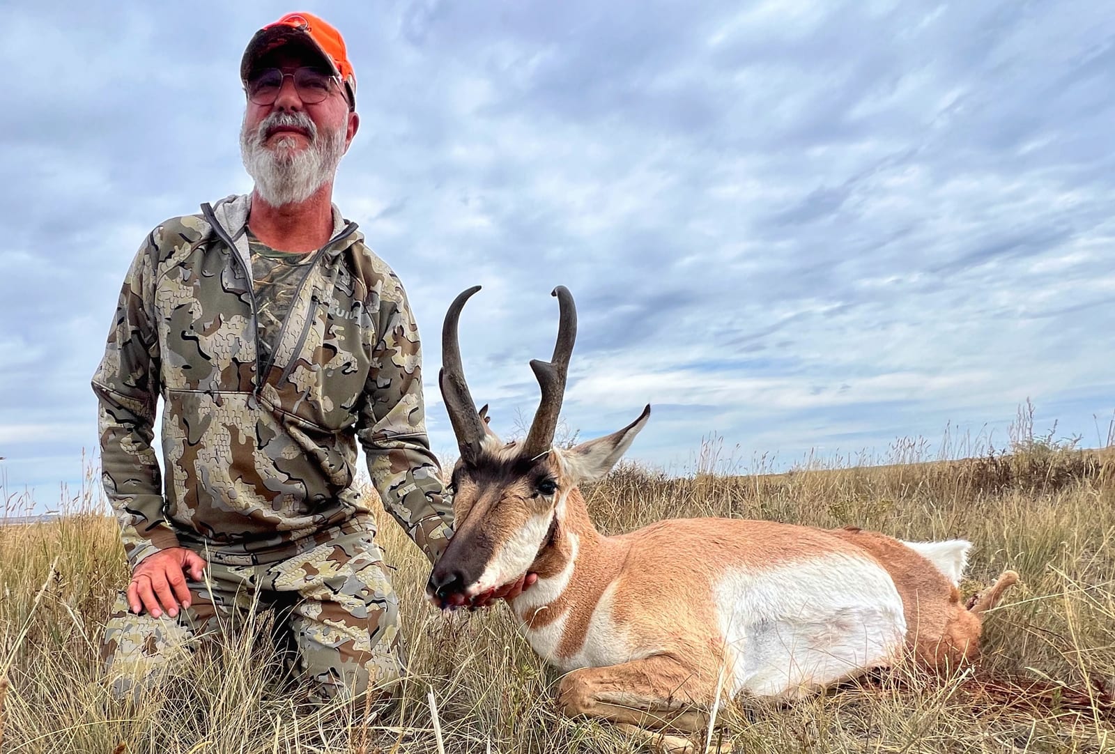 A hunter kneels next to a harvested pronghorn in a grassy field under a cloudy sky.