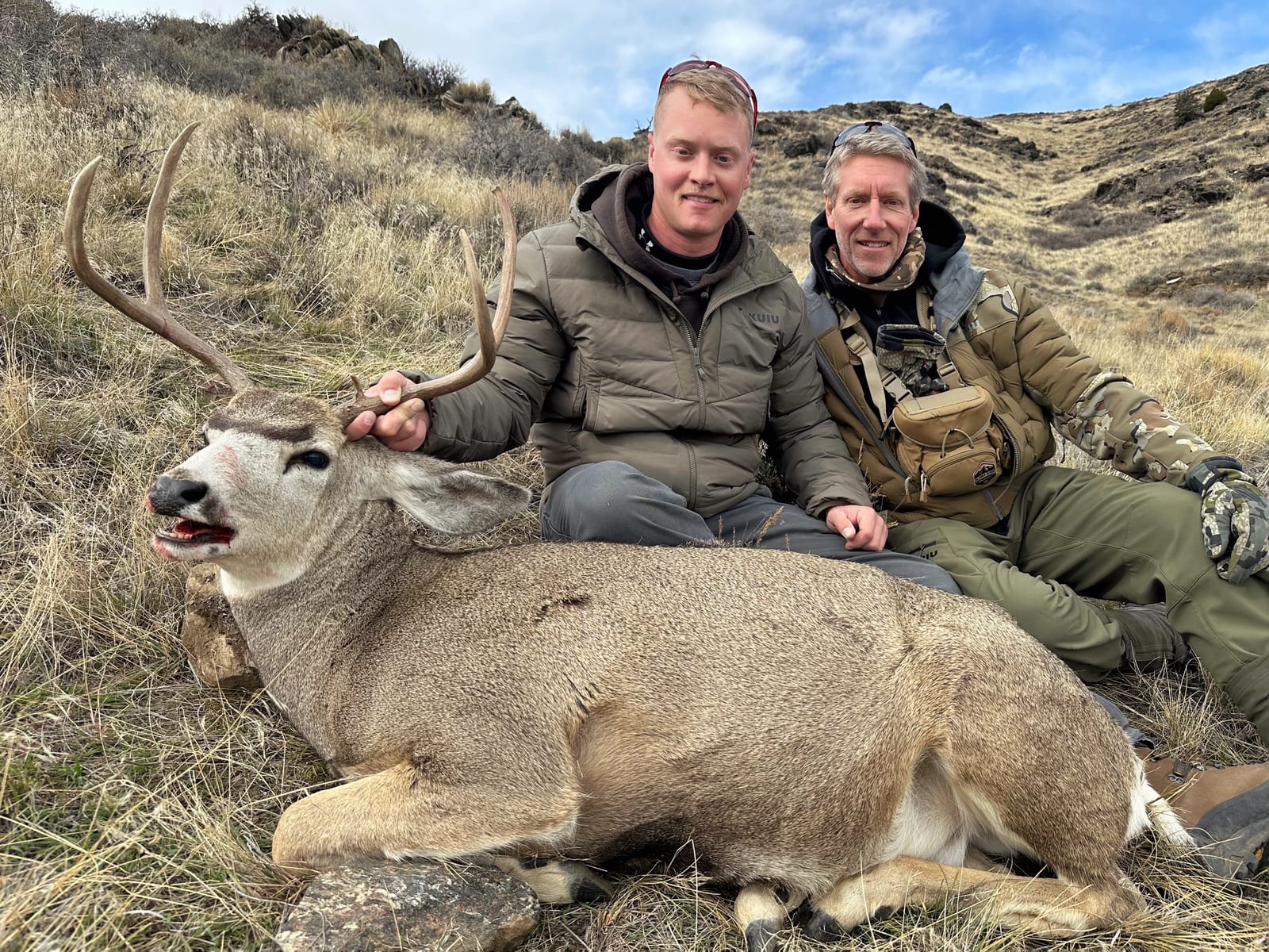 Two hunters pose with a harvested mule deer on a grassy hillside.