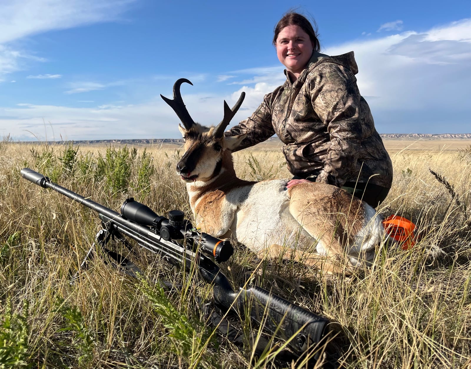 A woman in camouflage poses with a harvested pronghorn in a grassy field, with a rifle in the foreground.