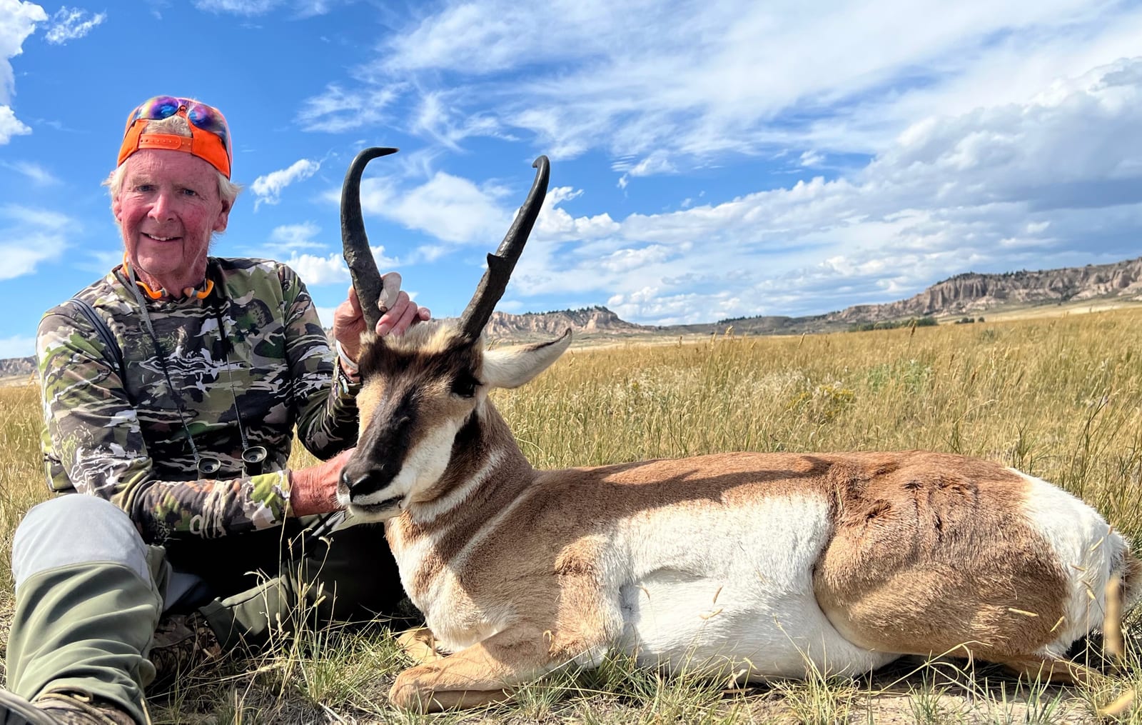 A hunter poses with a harvested pronghorn in a field under a blue sky with clouds.