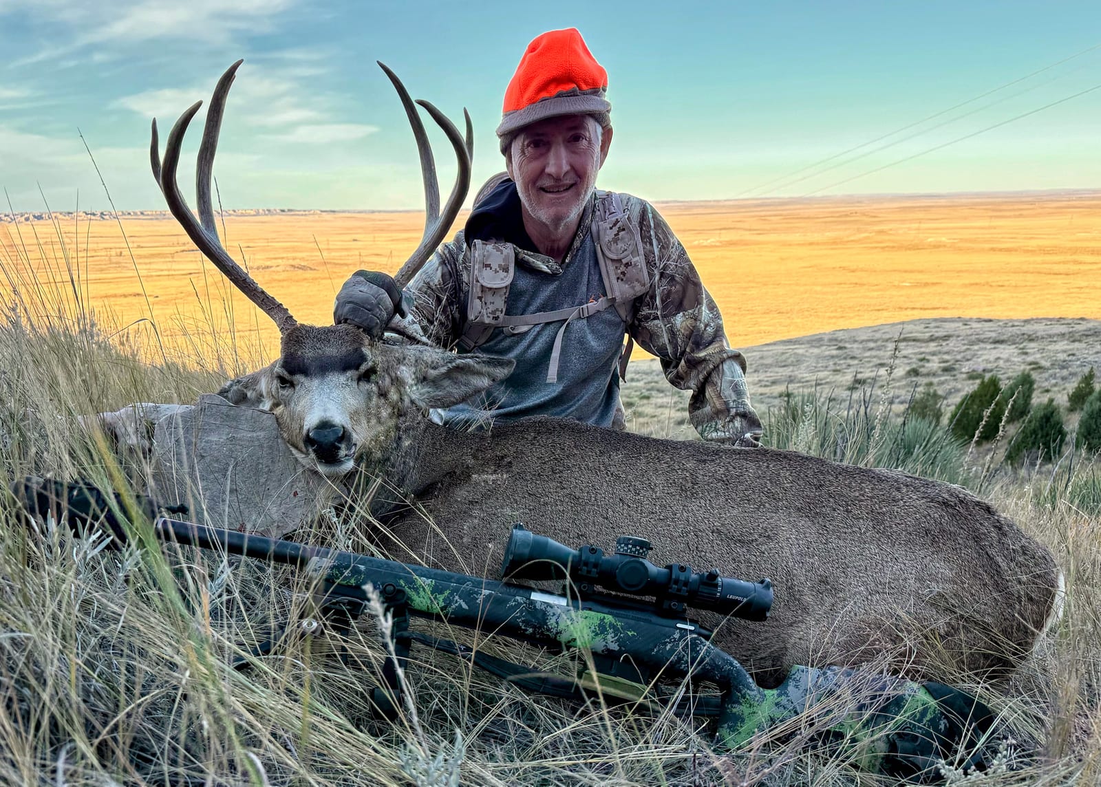 A hunter in an orange hat poses with a harvested mule deer and a rifle in a grassy field.