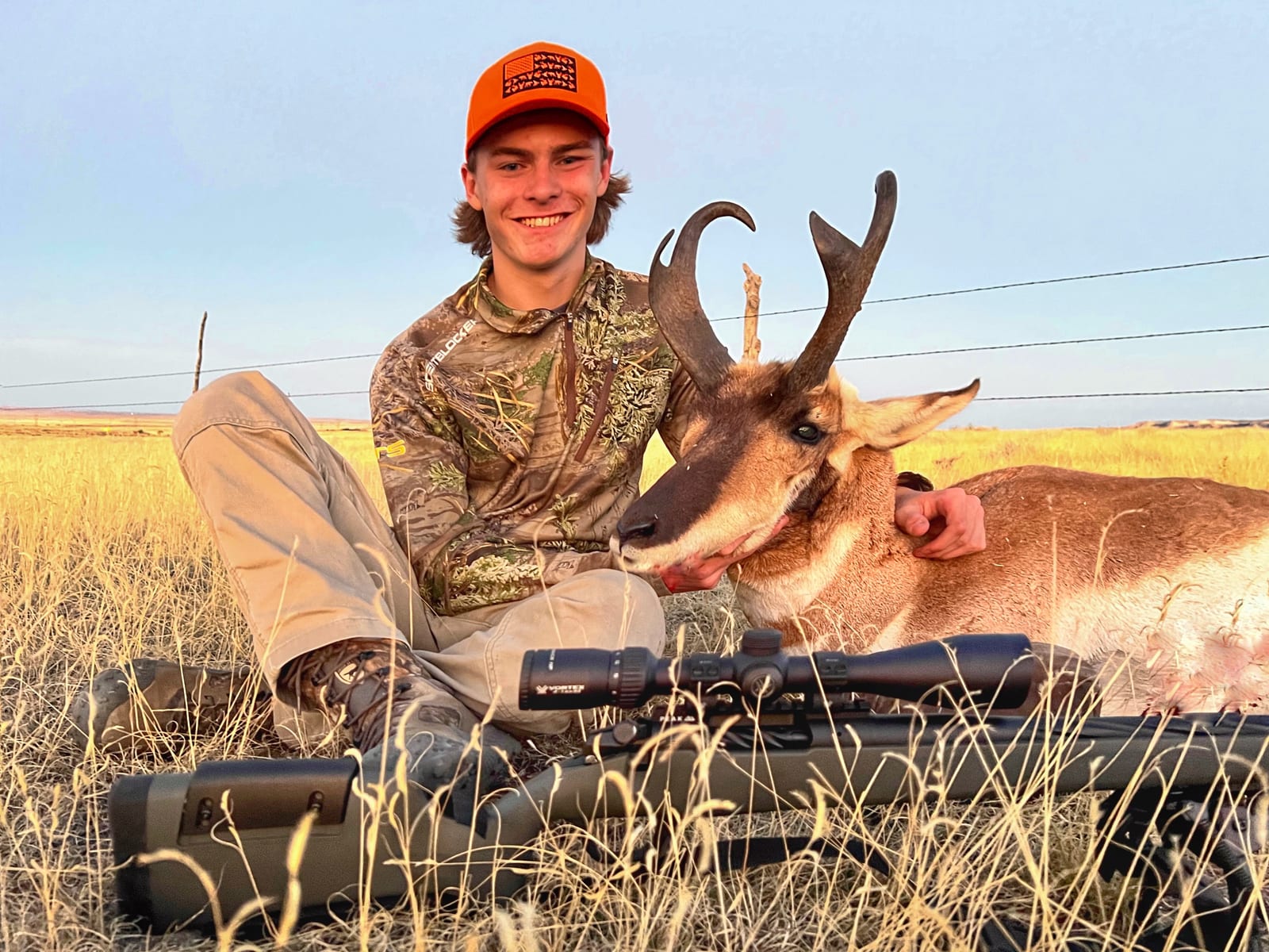 A young man in camouflage and an orange hat poses with a harvested pronghorn in a grassy field.
