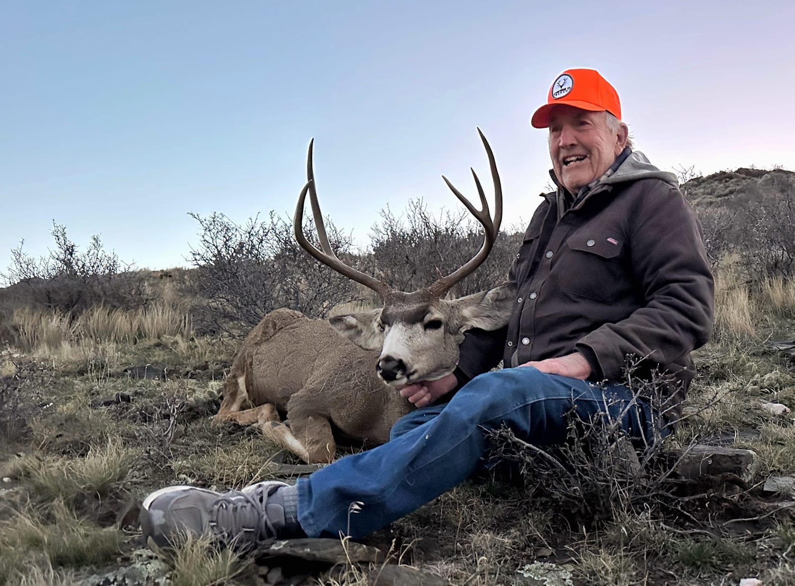 A hunter sits next to a harvested mule deer in a field.