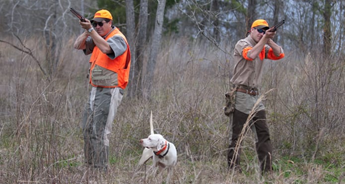 Two hunters with shotguns and a white hunting dog are in a field with dry grass and trees in the background.