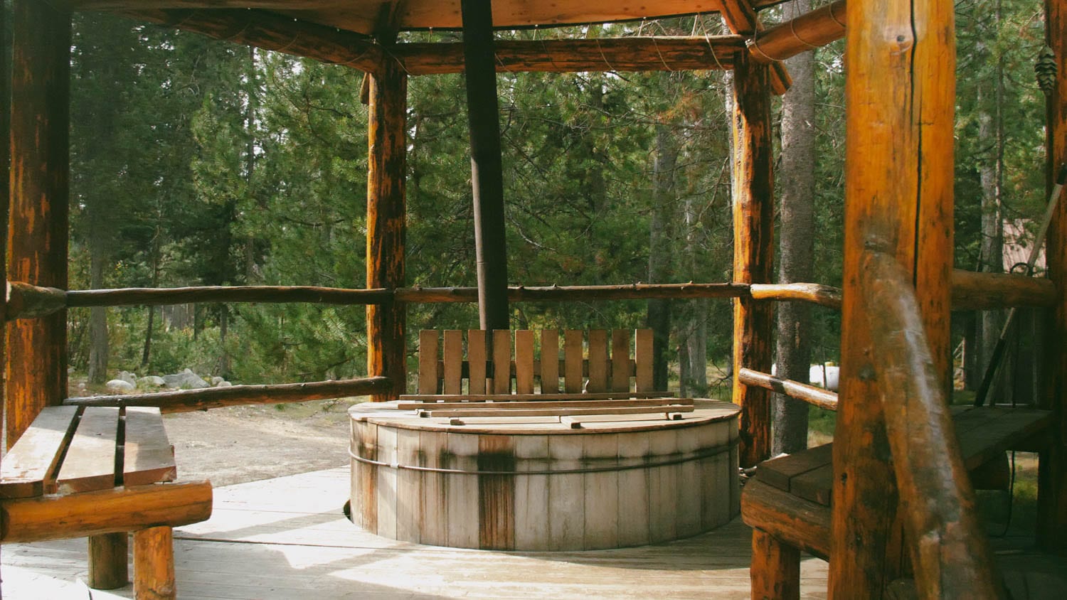 A wooden hot tub sits within a rustic log structure, surrounded by trees.