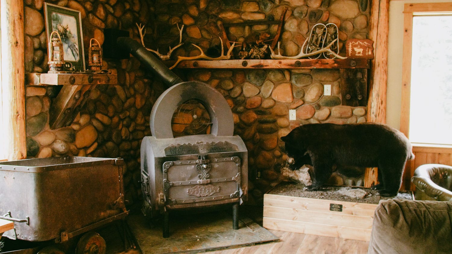 Interior view of a rustic lodge featuring a stone fireplace, a wood stove, and a taxidermied bear.