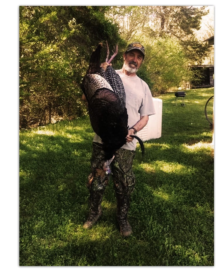A man poses with a harvested wild turkey in a grassy area.