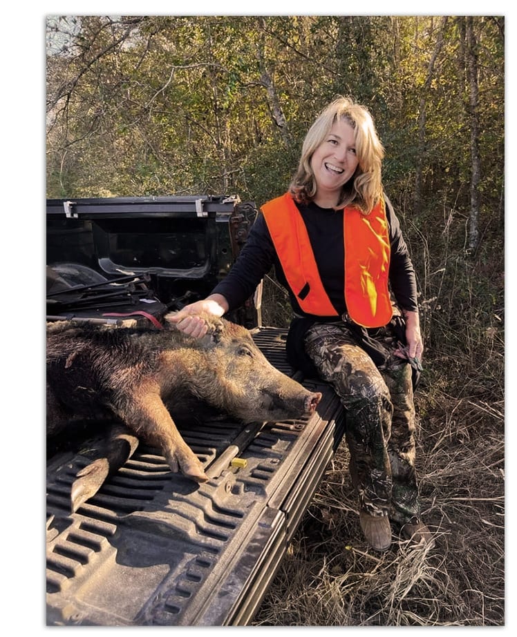 A woman in an orange vest poses with a harvested wild boar in the back of a truck.