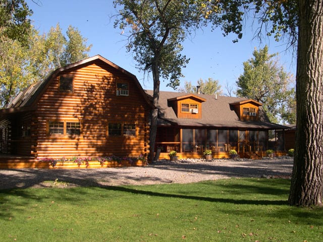 A large wooden lodge building with a green lawn in front and trees surrounding it under a clear sky.