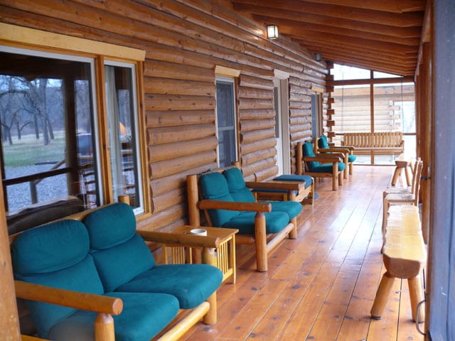 A wooden lodge porch features teal cushioned chairs and a wooden swing.