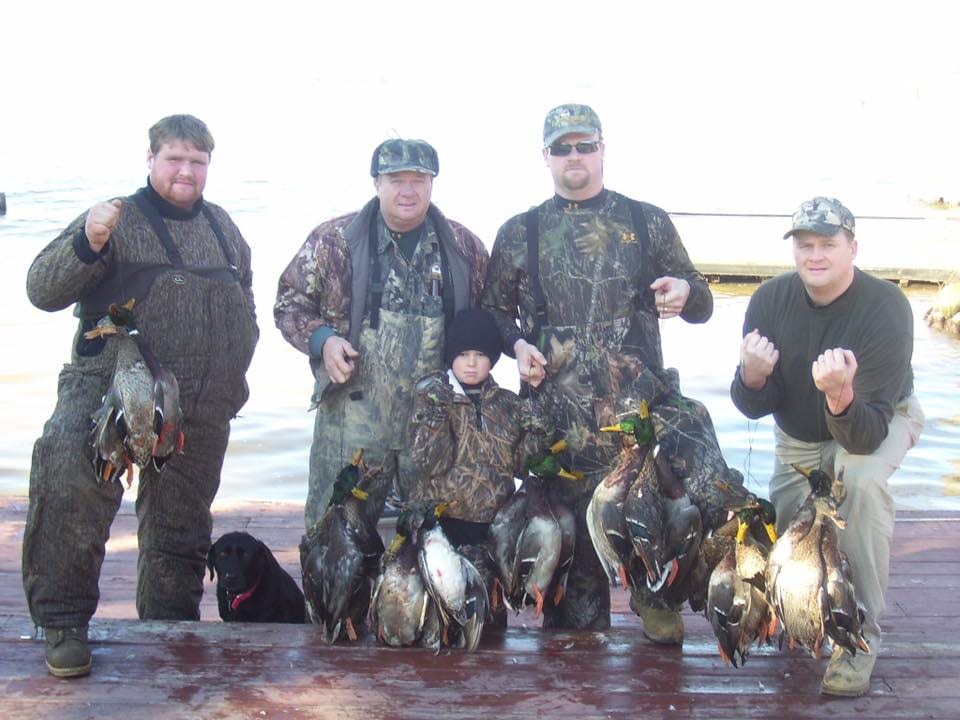 Several hunters and a child pose with their harvested ducks and a black dog on a wooden dock.