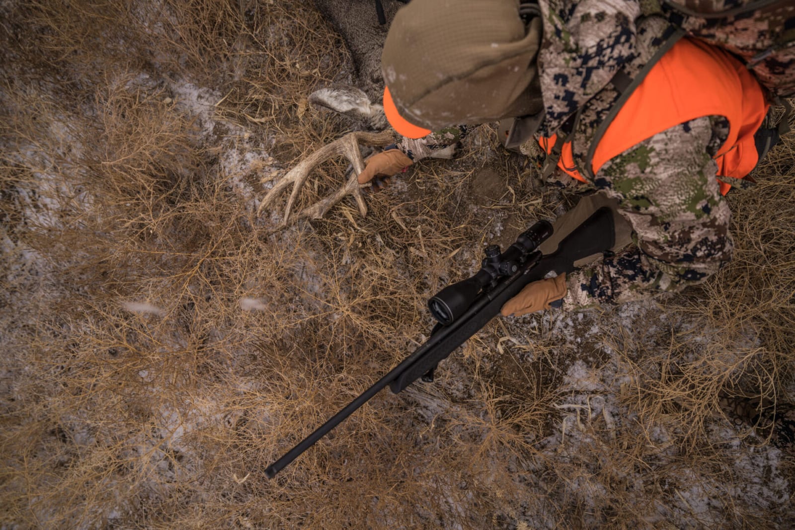 A hunter in camouflage and an orange vest holds a harvested deer antler on the ground next to a rifle.