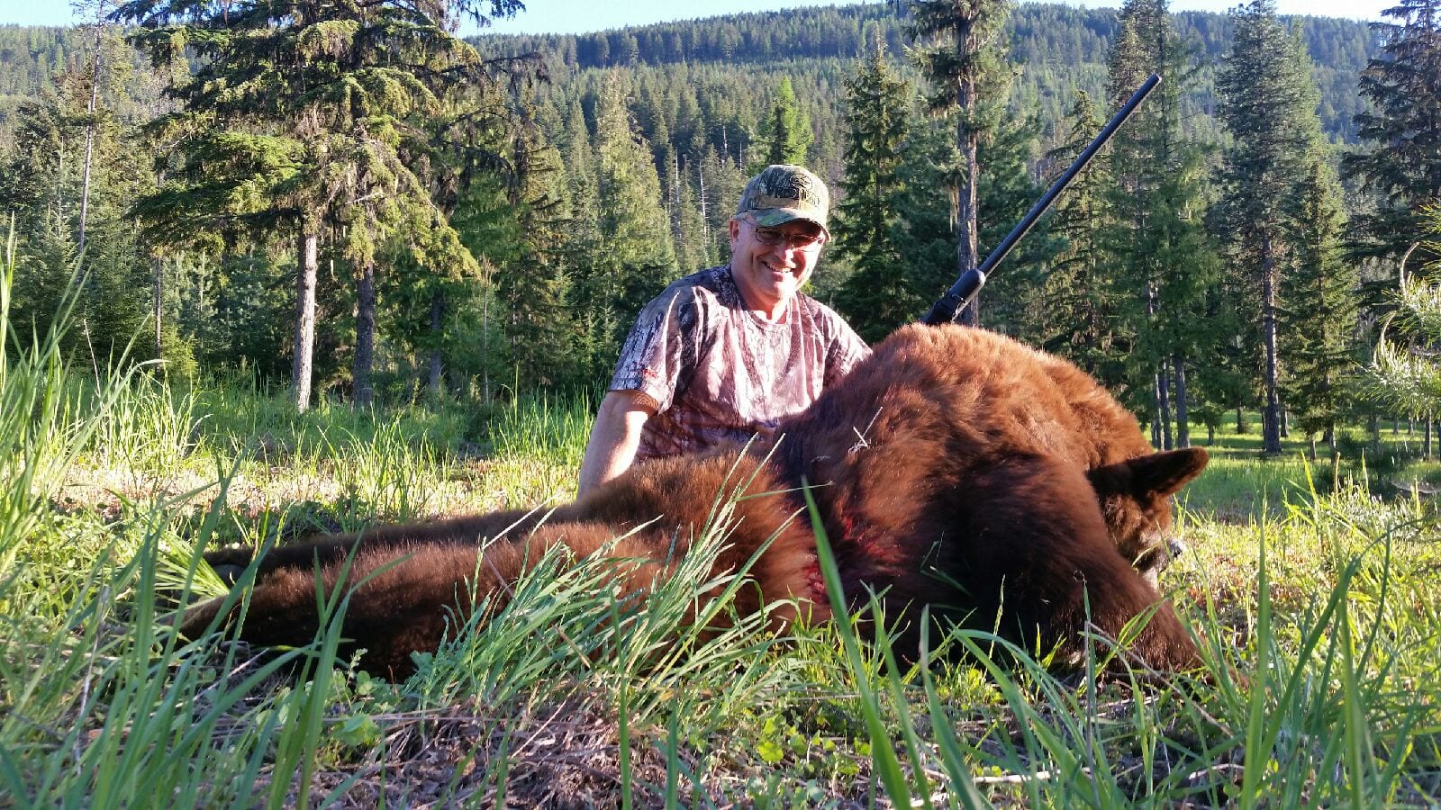 A hunter poses with a harvested black bear in a grassy, forested area.