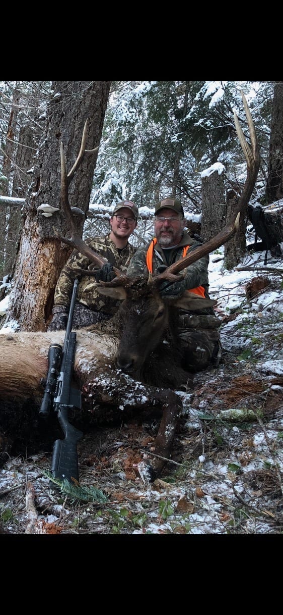 Two hunters pose with a harvested elk in a snowy forest.