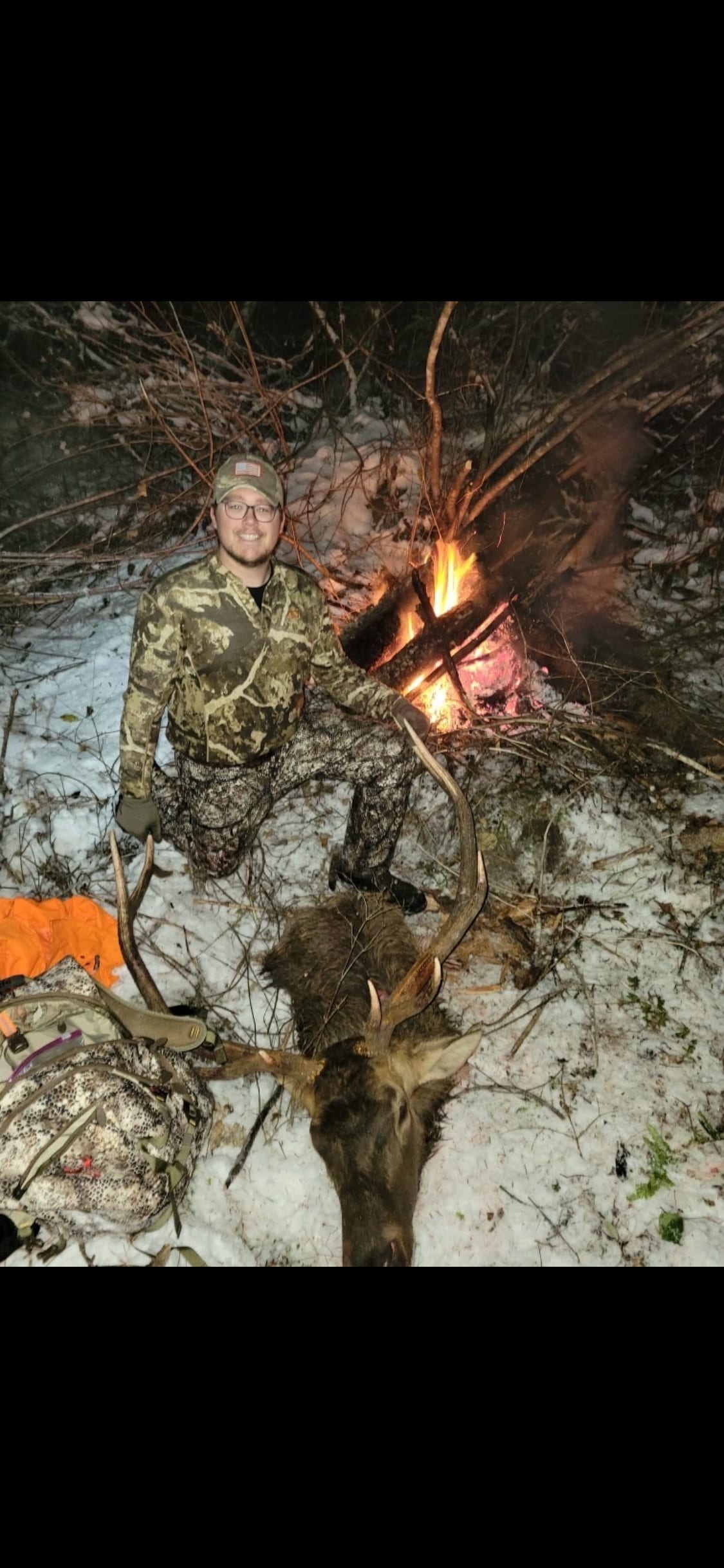 A hunter in camouflage poses with a harvested elk next to a campfire in a snowy, wooded area at night.