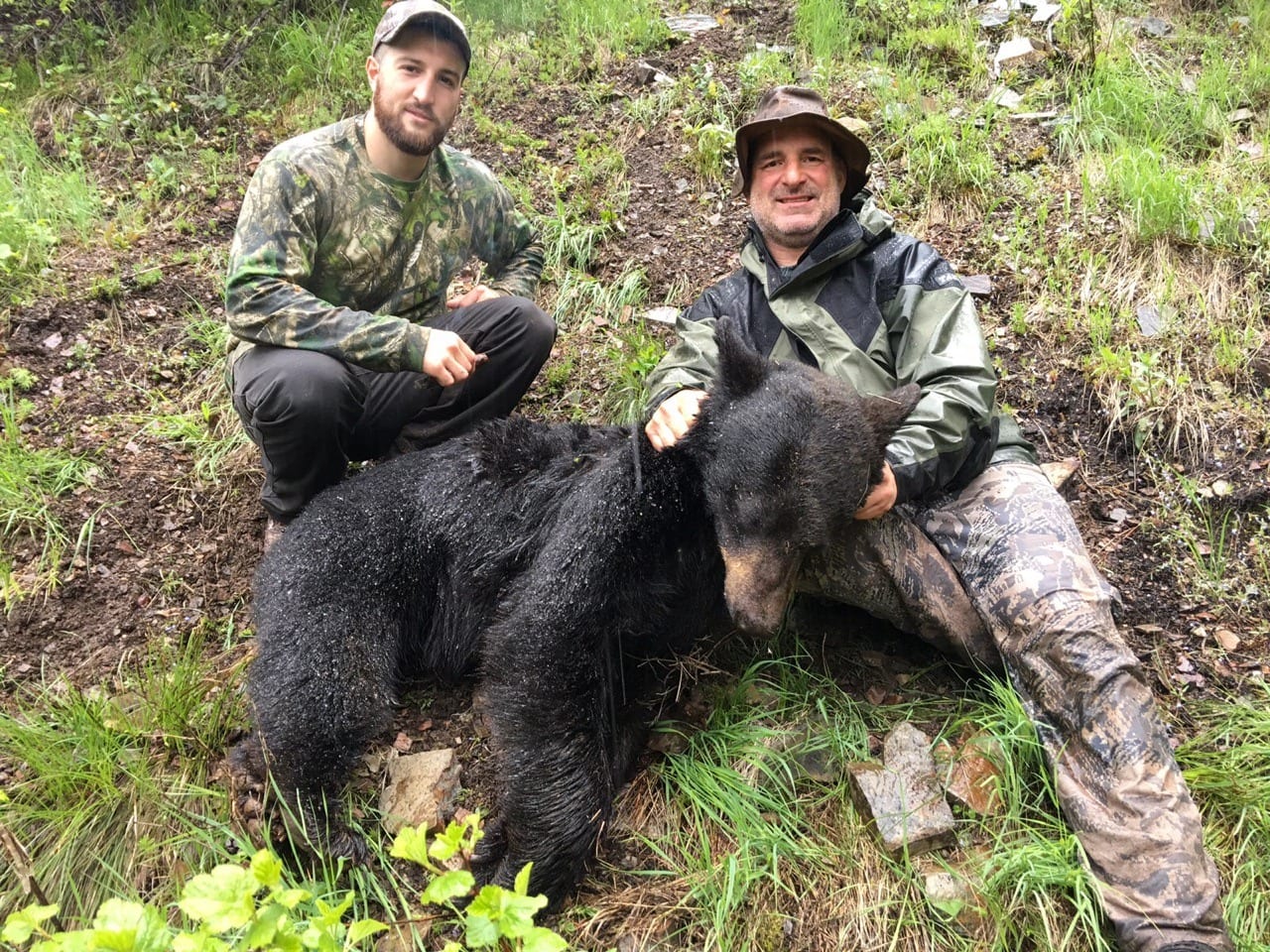 Two hunters pose with a harvested black bear on a grassy hillside.