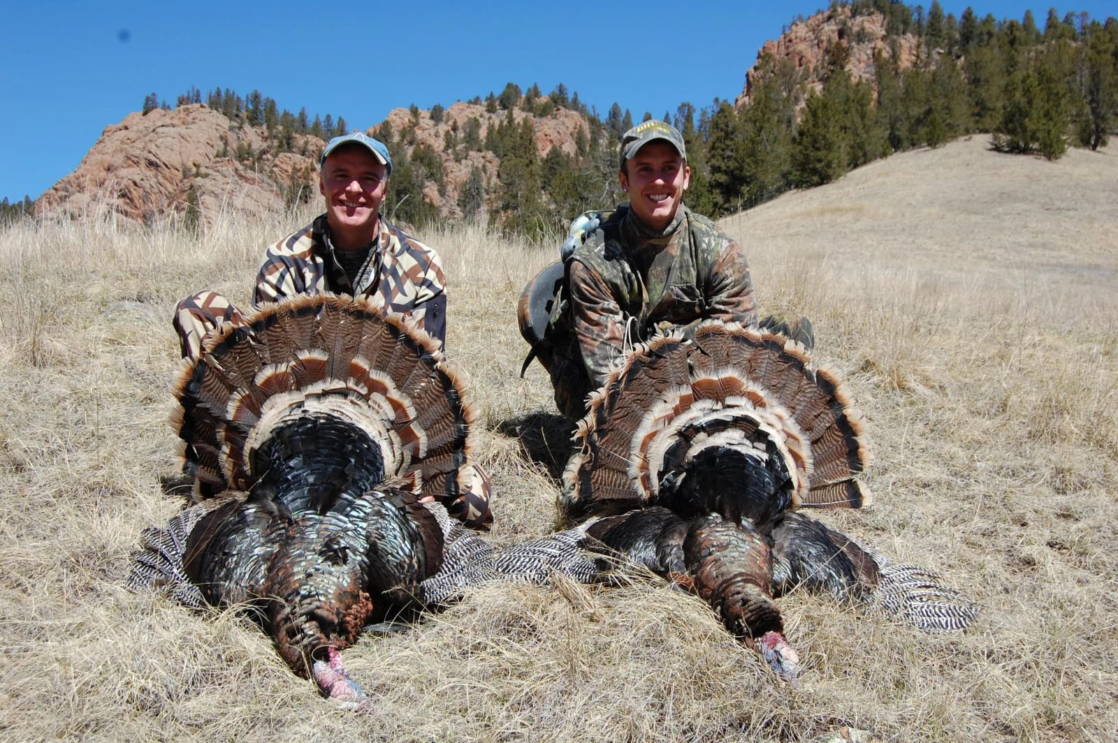 Two hunters crouch in dry grass with proud smiles, posing behind two large wild turkeys they hunted, with rocky hills and a partly cloudy blue sky in the background.