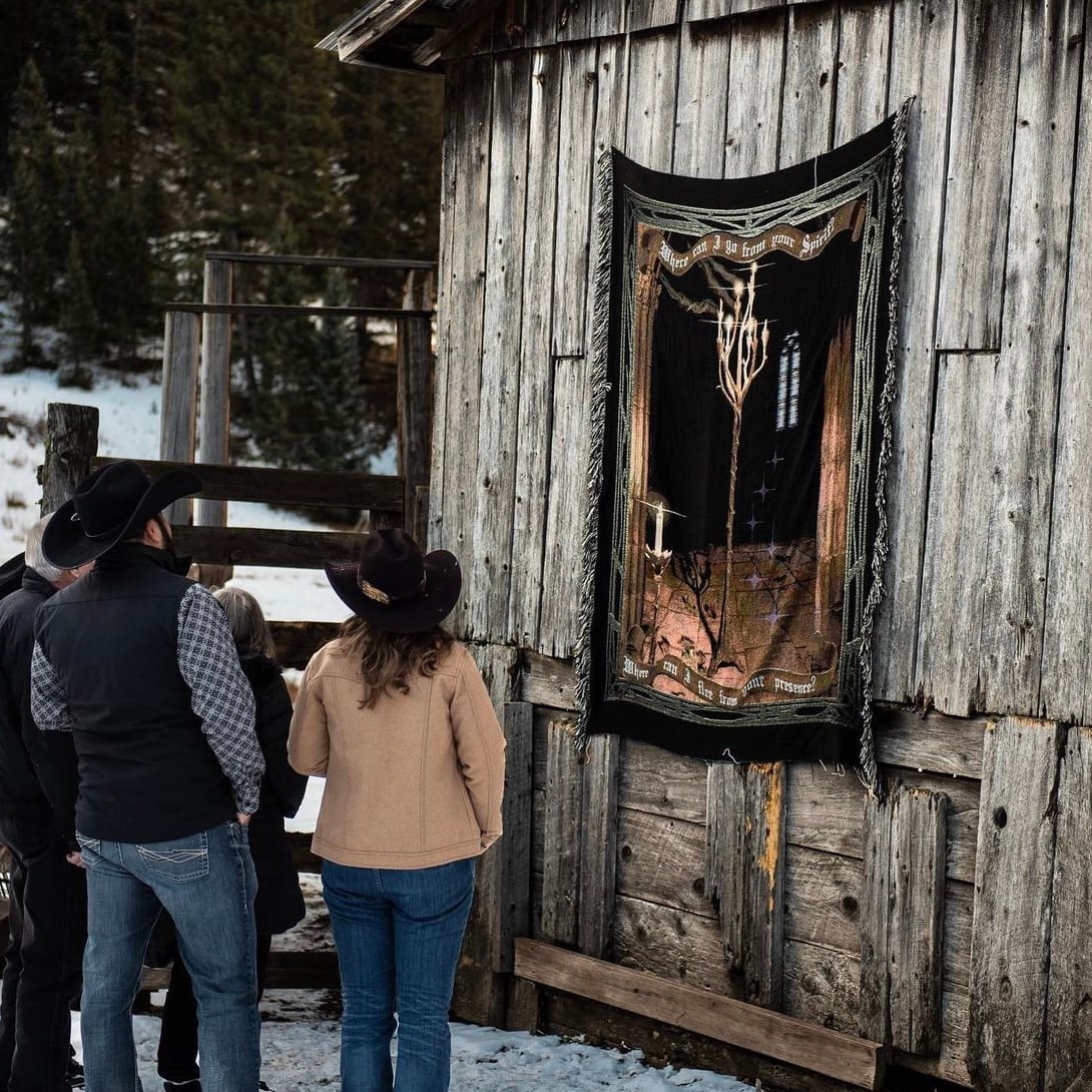 People in cowboy hats stand outside a rustic wooden building with a tapestry hanging on its wall, in a snowy, wooded environment.