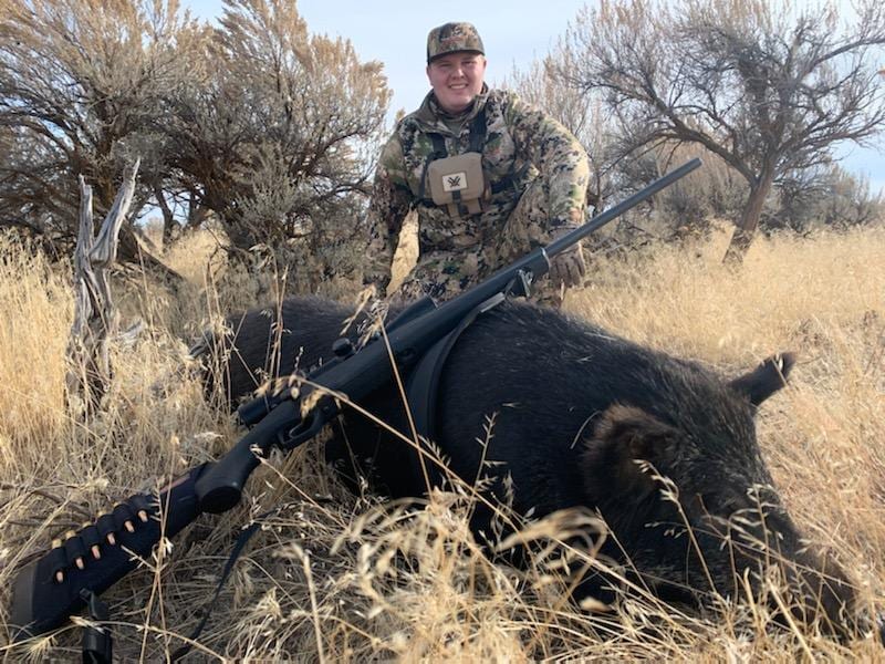 A hunter in camouflage poses with a harvested wild boar and a rifle in a grassy field.