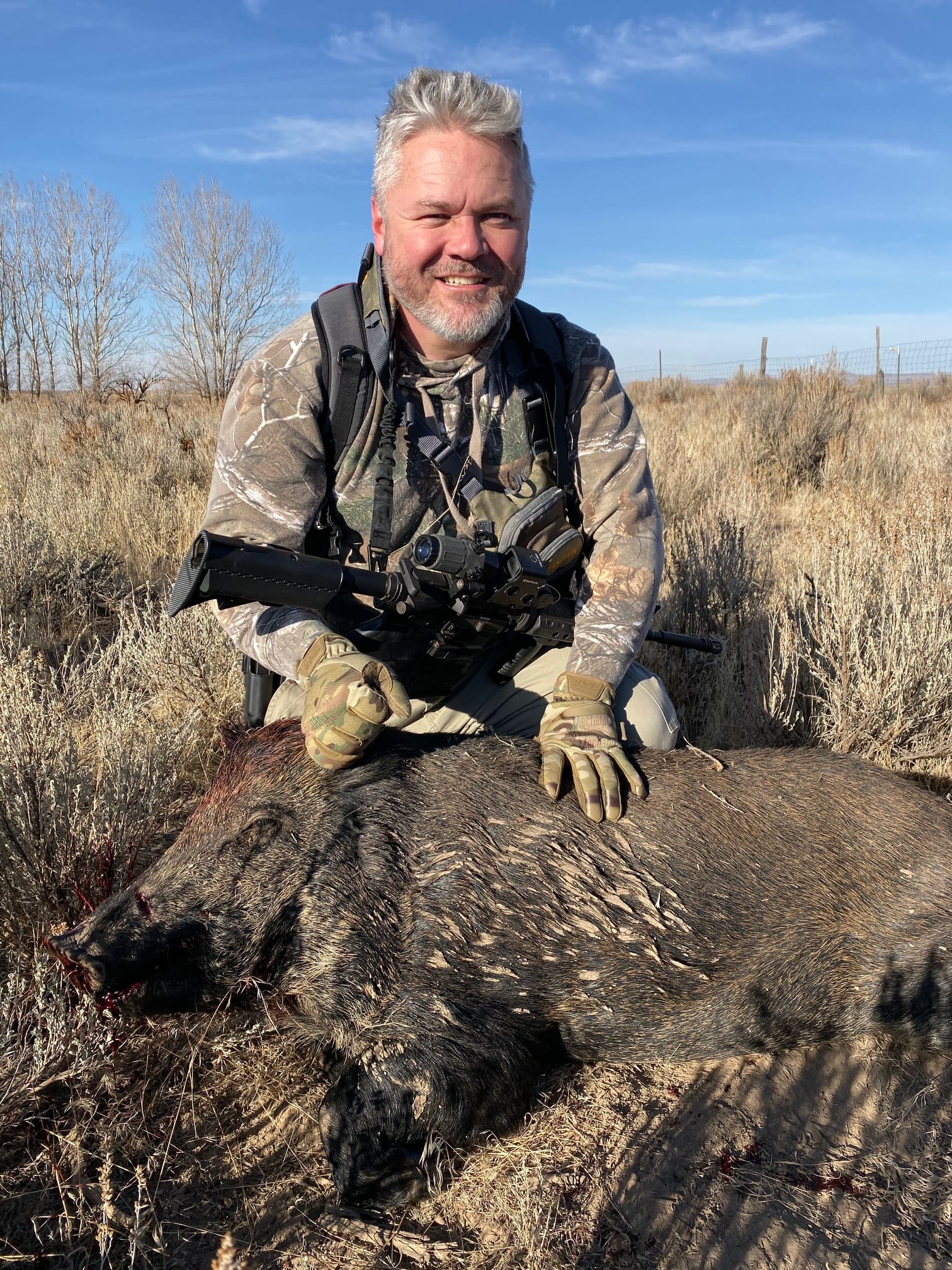 A man in camouflage poses with a harvested wild boar in a dry, grassy field under a clear sky.
