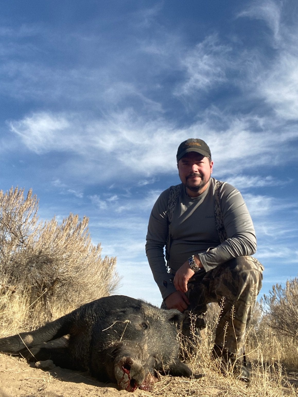 A hunter kneels beside a harvested wild boar in a dry, brushy field under a blue sky.
