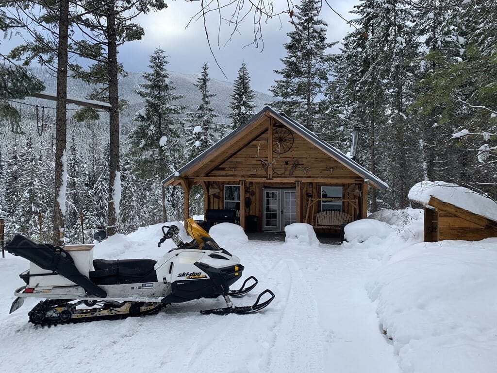 A wooden cabin in a snowy forest with a snowmobile parked in front.