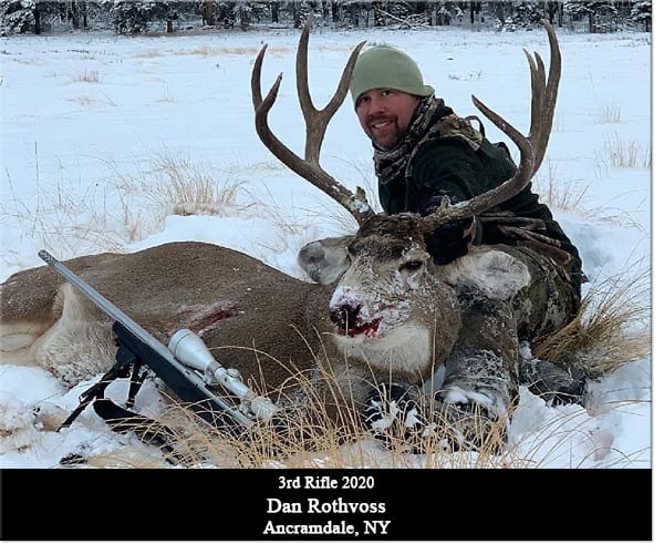 A hunter poses with a harvested mule deer in a snowy field.